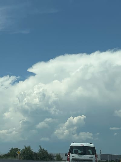 The image captures an outdoor scene in Tsaratsovo, Plovdiv Province, Bulgaria, under a partially cloudy sky during daylight hours. The upper two-thirds of the frame are dominated by a deep blue sky featuring large, bright white cumulus clouds, with one prominent cloud formation on the left exhibiting a distinct avian or anvil-like profile. In the lower portion of the image, a white multi-purpose vehicle (MPV) or small van is observed from the rear, positioned on a road. The vehicle is equipped with roof rails, and its license plate partially displays "PB," corresponding to the Plovdiv region. To the left of the vehicle, green foliage comprising trees and bushes is visible, with a yellow diamond-shaped road sign partially obscured within the vegetation. In the background, beyond the immediate roadside elements, several large, grey, rectilinear industrial or warehouse buildings stretch horizontally, indicating a commercial or industrial zone in the distance. No individuals are discernible within the frame.
