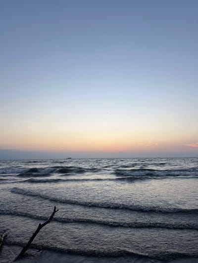 A horizon line divides a landscape into sky and sea. The left portion of the image is occupied by a gradient sky, transitioning from pale blue to a golden hue near the horizon. To the right, waves roll rhythmically, reflecting the light in shades of grey and silver. A dark branch protrudes into the lower right corner of the sea, adding a natural element to the scene. A very small boat or structure appears on the sea near the horizon.