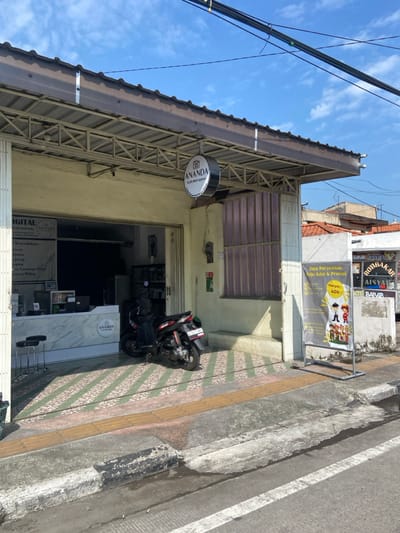 The image shows the exterior of a storefront in Kecamatan Tulangan, Indonesia, located at latitude: -7.482, longitude: 112.647. The storefront is a single-story building with a brown awning and a sign that reads "ANANDA" in white lettering. The awning is supported by a metal frame. There is a large window on the right side of the storefront, which is covered with a white plastic sheet. The front of the storefront is paved with a black and white checkered tile pattern. A black motorcycle is parked in front of the storefront, near a step leading up to the entrance. There is a black metal stool to the left of the entrance, along with a marble counter and a small sign that reads "ANANDA". A sign for a clothing store advertising a 40% sale is located on the right side of the storefront. The building next door is visible behind the sign, with a sign in Indonesian reading "ROTI BAKAR Aisyah" in white lettering. The street in front of the storefront is paved with asphalt and a white stripe is visible on the right side of the image. The sky above the street is blue and partly cloudy.