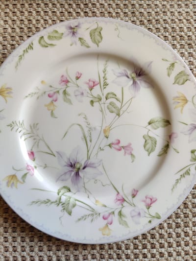 A white ceramic plate with a floral pattern is placed on a textured, woven brown placemat. The plate features delicate watercolor-style paintings of purple clematis, pink sweet peas, and yellow bell-shaped flowers, interspersed with green leaves and tendrils. A light lavender scalloped border encircles the rim of the plate. The scene is photographed from a top-down perspective. This image is not associated with the city of Rybnitsa, Moldova, as there are no elements in the image to suggest a specific geographical location.