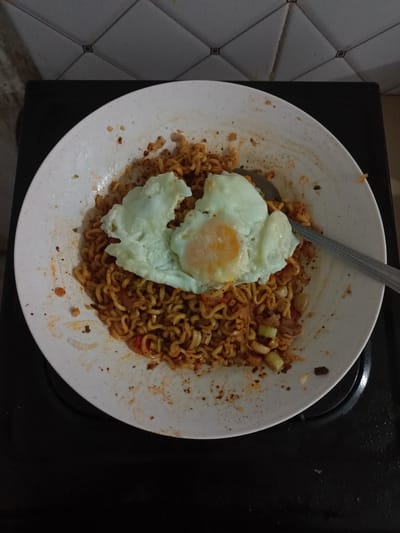 A top-down shot depicts a white plate filled with noodles and a fried egg on a black surface, presumably a stove top. A metal spoon rests on the plate amidst the noodles. The setting appears to be a kitchen, evidenced by the tiled wall partially visible in the background. Given the prompt, it is implied this scene is located in Batam, Indonesia. No people are visible.