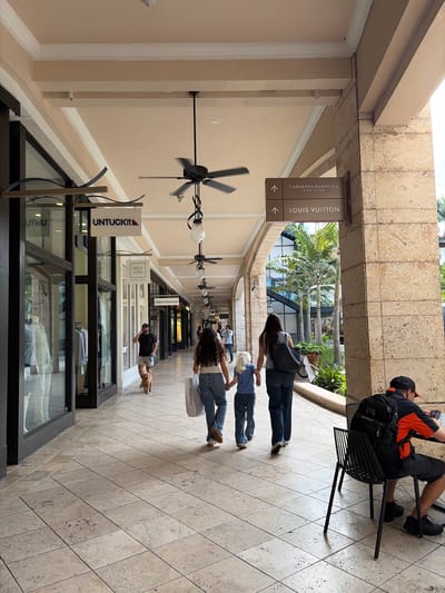 The photo shows a long corridor in what appears to be an outdoor shopping mall in Coral Gables, United States of America. There are retail storefronts on either side of the walkway. Signs for stores such as "UNTUCKit", "120% Lino", and "Allen Edmonds" are visible, along with a sign pointing to "Carolina Herrera" and "Louis Vuitton" in one direction.
Several people are walking along the corridor, including a family of three (two adults and a child), a man with a dog, and others in the distance. There is a man sitting on a chair near the entrance to the corridor, possibly using a phone.
The architecture features archways and decorative lighting fixtures. The floor is tiled with light-colored stone. There are large ceiling fans along the corridor.