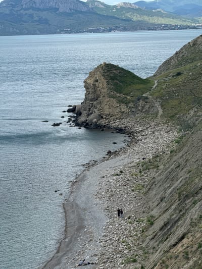 The image depicts a coastal landscape, possibly near Орджоникидзевский поселковый совет, Ukraine. A rocky coastline slopes down to a body of water. A small promontory extends into the water.  Two figures are present on the shore. The area is rocky and partly vegetated. Mountains are visible in the distance.