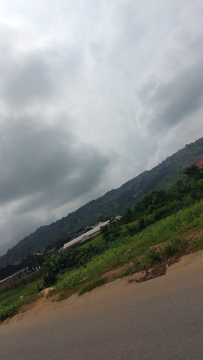 The image depicts an angled view of a sloping hillside covered in vegetation and scattered buildings, suggesting a semi-urban or rural setting. A paved road runs diagonally across the foreground, and the sky above is overcast with heavy, grey clouds. The hillside features dense green foliage interspersed with several structures, some appearing to be residential houses with white roofs and walls, and others possibly commercial buildings. The vegetation ranges from low-lying crops to taller trees and shrubs. In the mid-ground, a few people are visible, including two individuals on what appears to be a motorcycle, near some of the buildings. The overall impression is of a landscape in Bwari, Nigeria, characterized by natural topography and human development.