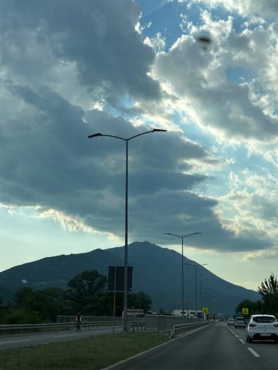 The image shows a view from a vehicle driving on a highway in Begovina, Montenegro, with a large mountain range in the background under a dramatic sky filled with dark clouds and shafts of sunlight. In the foreground, the road curves slightly to the right, with several cars visible. A white hatchback is in the right lane. On the left side of the road, a cyclist wearing a red and white jersey is riding. A bridge with a metal railing is visible parallel to the highway. Several tall streetlights with double arms are spaced along the roadside. In the middle distance, a dark, square signpost stands near the edge of the road. The mountain range dominates the horizon, appearing dark and textured, with trees and some structures visible at its base. The sky is a mix of dark, heavy clouds and brighter, sunlit patches, creating a sense of depth and dynamic weather.