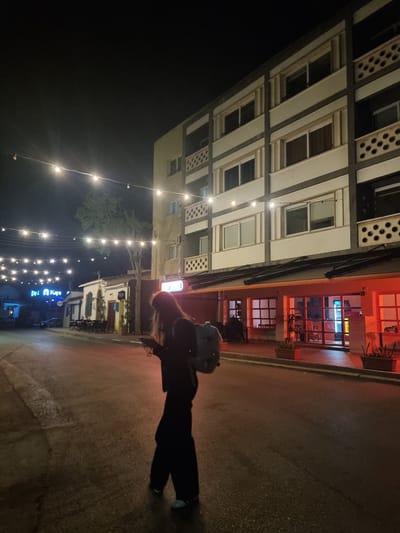 A photograph taken at night in North Nicosia, Northern Cyprus, shows a person walking along a street. The person is wearing dark clothing and carrying a backpack. To the left, a multistory building with regularly spaced windows is visible. A cafe or bar with an orange facade is next to the building. Overhead string lights extend down the street, illuminating shops and businesses.