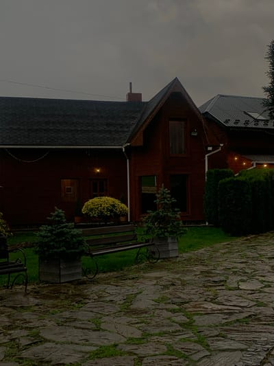 The scene, located in Lazeshchyna, Ukraine, depicts the exterior of a multi-story building under a dark, overcast sky, with visibly damp ground surfaces. The primary structure is a dark reddish-brown wooden building featuring a dark shingle roof. A red brick chimney is positioned on the left roof section, while the right section presents a steep gable with a tall, narrow window. An adjacent building section to the right has a dark metal roof with visible vents. The building's facade includes a small square window, a rectangular window, and a door on the lower left; a single light fixture is illuminated above this door, and string lights are strung horizontally across the front and along the gable. The foreground consists of an irregular flagstone pathway with grass growing between the stones. A strip of green grass separates the pathway from the building. On this grass strip, two dark wooden benches with decorative black metal supports are positioned, each flanked by a small evergreen tree in a square wooden planter. A large, spherical, yellow-leafed shrub in a pot is situated near the center of the building's facade. A dense line of tall, dark green hedges forms a boundary along the right side of the frame. No individuals are present, and no actions are depicted.