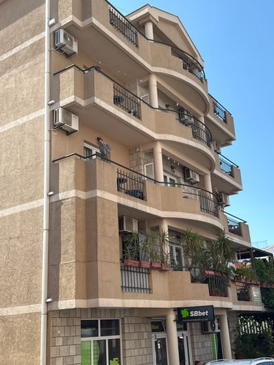 This image captures a close-up, low-angle view of a multi-story building with a light brown stucco facade, situated in Lapčići, Montenegro. The building features multiple curved balconies with black metal railings. Several air conditioning units are visible mounted on the exterior walls of the building.

On one of the balconies, a person, partially obscured, is standing near a window. A gray garment is hanging on a clothesline stretched across the balcony. Plants in rectangular planters are arranged along some of the balconies.

At the base of the building, there is a brick facade with several storefronts. One prominent storefront features a sign with the logo "SBbet SPORTING BOOKMAKERS / est. 1997". Another sign, partially visible, also appears to be related to "SBbet". A white car is parked in front of the building. The sky above is clear and blue. The overall impression is one of a commercial and residential building in an urban or semi-urban setting.