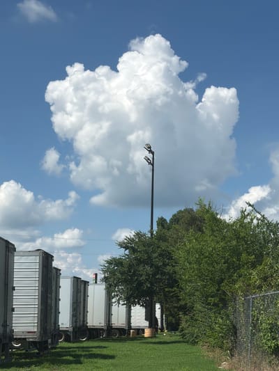 The image shows a line of parked semi-trailer trucks on a sunny day in Lebanon, United States of America. The trucks are silver and white, with visible tires and landing gear. They are parked on a grassy area next to a chain-link fence. Behind the trucks, there are various trees and bushes. A tall pole with two lights is also visible in the background, along with a large, fluffy white cloud in a clear blue sky. The scene appears to be an outdoor storage or parking lot, possibly near a distribution center or industrial area.
