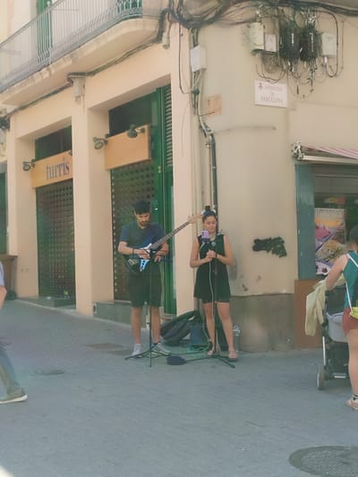 A photograph shows two street musicians performing on a street corner in Rubí, Spain. A man with dark hair and a beard is playing an electric bass guitar. A woman with dark hair is singing into a microphone, holding a cell phone. They stand in front of a building. The building features closed storefronts with the word "Turris" above one, and the number "2" next to the other. An "Avinguda de Barcelona" street sign is visible above a building corner. Overhead electrical wires and junction boxes are grouped near the sign. To the right, a woman is pushing a stroller.