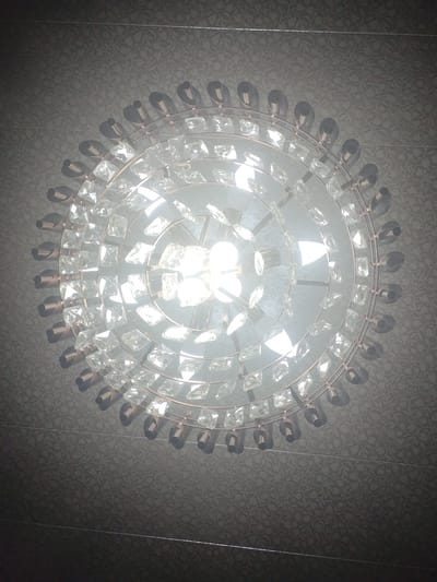 This is a close-up, top-down photograph of a modern chandelier. The chandelier is circular and features multiple tiers of clear, faceted crystal pendants. These crystals are suspended from thin, copper-toned metal rings and wires. The central part of the chandelier contains bright, exposed light bulbs that are emitting a strong glare, obscuring some of the inner details. The chandelier is hanging from a ceiling that has a dark, textured wallpaper with a subtle floral or leaf pattern. There are no people or other objects visible in the image. The location is not discernible from the image itself.