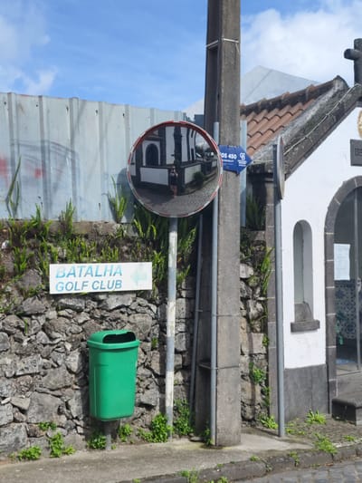 The image captures a street scene in Ponta Delgada, Portugal. A weathered stone wall covered with ferns and plants forms the backdrop, with a sign indicating "Batalha Golf Club" pointing left. A green trash can is mounted to the wall's left side. A convex mirror is mounted on a pole that has a utility pole behind it, reflecting an adjacent building and a woman. Adjacent to the pole, a small white chapel with a tiled roof and a cross on top is visible. A Coldwell Banker sign is fixed to the utility pole near the chapel. The street is paved with cobblestones. A corrugated metal fence is positioned behind the wall. The sky is blue with scattered clouds.