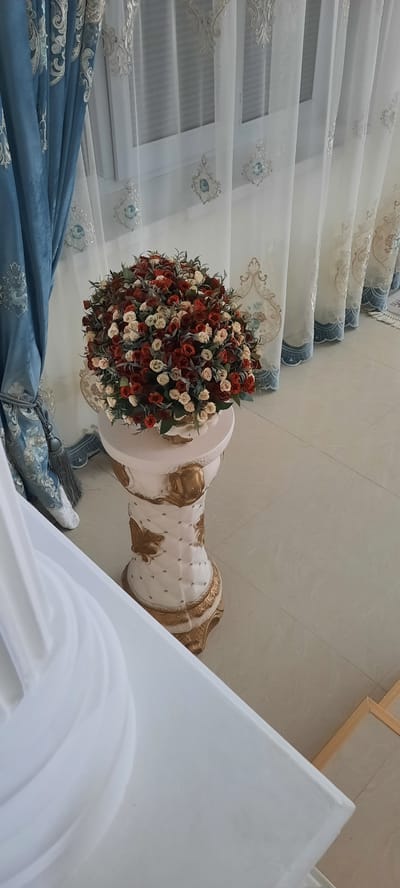 The image depicts an interior scene. In the foreground, a white draped object occupies the lower left portion. To the right of this, a decorative pedestal stand is visible, featuring intricate gold detailing and a quilted pattern. Atop the pedestal is a large, spherical arrangement of artificial flowers, predominantly in shades of red and cream, interspersed with greenery. Behind the pedestal and flowers, elegant curtains are displayed. On the left, a heavy, dark blue curtain with ornate embroidery is partially drawn. To the right, sheer white curtains with embroidered accents are visible, revealing a window behind them. The floor is tiled in a light beige color. The scene suggests a well-decorated interior space.