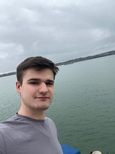 A male individual is prominently featured in a waist-up selfie, positioned on the left side of the frame. He is a young adult with short dark brown hair, fair skin, and light stubble on his chin and jawline. He wears a plain grey short-sleeved t-shirt and looks directly at the camera with a slight smile. Behind him, a large body of choppy, greenish-blue water extends into the distance under a uniformly grey, overcast sky. In the mid-ground, a long, dark-colored cargo ship or barge is visible on the water, positioned horizontally relative to the frame. Further in the background, a hilly coastline with green vegetation and scattered light-colored buildings is visible across the water. In the bottom right corner of the foreground, a blue, ribbed surface is partially visible next to a circular yellow object displaying black text and symbols. This scene depicts a view over the Menai Strait from Bangor, Wales, United Kingdom.