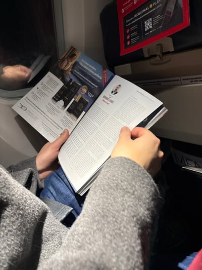 A person is shown from an overhead perspective, reading a magazine inside what appears to be an airplane or similar public transport. The individual's hands are holding the open magazine, with a gray-patterned jacket or coat visible on their arm and lap, covering blue jeans or trousers.
The magazine features an article on the right page with a small portrait of a man, likely the author or subject, and the title "REFORMANDO A BOBOT." The left page displays an advertisement for "Pompeia Life" products, titled "Próximo destino: Una salud íntima perfecta," showcasing product bottles and steps labeled "Paso 1 Step" and "Paso 2 Sep 2." The ad includes the text "De venta en www.pompeia.es/ritual, envío Internacional disponible."
In the background, to the left, a dark seat pocket or storage area is visible, containing a red-bordered card or screen that reads "Descubre REGIONAL PLAY" and "Nuestra plataforma de entretenimiento a bordo" with a QR code. Above this, a light-colored panel or overhead compartment section displays text: "CHALECO SALVAVIDAS," "LIFE VEST UN," and "METRAS PERMANEZCA SENTADO." Part of the word "MEN" is also visible, possibly from "MENU" or a similar sign. A blurry reflection of a hand is seen in a dark, possibly window-like surface below the magazine. The overall lighting is dim, suggesting an indoor setting, possibly during the evening or night. The presence of "LIFE VEST" and "in-flight entertainment" strongly indicates an airplane setting.