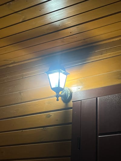 A vintage-style outdoor lantern is mounted on a wooden wall, illuminating a portion of the exterior of a building. The lantern has a dark metal frame and a clear glass enclosure, with a bright light emanating from within. Below the lantern, a dark-colored door or cabinet is partially visible. The wooden wall is constructed of horizontal planks with visible grain and texture. The scene appears to be at night or in a dimly lit area, given the illumination provided by the lantern. The location is specified as Vyshhorod, Ukraine.