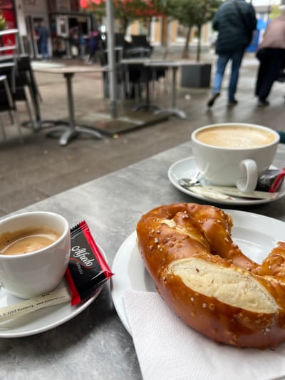 The image captures a scene at a cafe in Kehl, Germany. A pretzel rests on a white plate with a napkin. Beside it are two cups of coffee, each with a saucer and a small package of Alfredo Espresso coffee and a sugar stick. The table is outdoors, with a grey surface. In the background, blurred figures of pedestrians are visible on a sidewalk, along with buildings and trees characteristic of an urban environment. The day appears overcast, with soft, diffused light.