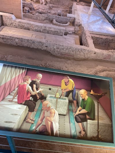 The image displays an archaeological excavation site in Alcalá de Henares, Spain, foregrounded by a detailed interpretive diorama. The background reveals exposed Roman ruins, including various-height stone foundation walls, earthen floors, and a partial white geometric mosaic. Modern metal support structures are visible on the upper right. The foreground diorama presents five stylized male figures within a simulated Roman interior, characterized by pink walls, a red-and-white striped wall section, and a light-colored floor with dark channels. The figures are positioned around stone benches containing circular latrine openings and a central rectangular stone basin. One figure, wearing a yellow tunic and blue trousers, stands over the basin, actively stirring its contents with two wooden implements. Two figures, dressed in red and dark tunics, are seated on a bench on the left, engaged in conversation. A bald, bearded figure in a white tunic squats beside the central basin. On the right, another bald figure in a green tunic is seated on a bench next to an ornate bronze-colored stand holding a flaming oil lamp.