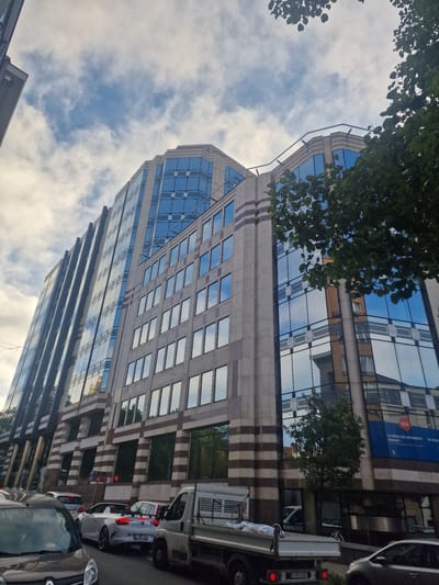 This is a ground-level, low-angle shot of a modern, multi-story office building with a glass facade reflecting the sky and surrounding buildings. The building has beige stone accents with dark horizontal stripes. In the foreground, there is a traffic jam on a street, with several cars visible, including a white convertible and a light-colored truck. Trees are present on the right side of the image, partially obscuring the building. The sky is partly cloudy with patches of blue visible. A blue banner with white text is attached to the building on the right side, displaying "LA RÉGIE DES BÂTIMENTS ENGAGE!" and "DE REGULIERE. A VOUS VOTRE COLLEGE !". This scene is located in Saint-Gilles - Sint-Gillis, Belgium.