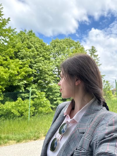 A young woman is captured from a low-angle perspective, seemingly looking down. She has brown hair styled with a braid visible at the back. She is wearing a gray plaid blazer over a light pink top with a bow detail at the collar. A pair of sunglasses is tucked into the blazer's lapel.
The background consists of lush green trees and a bright blue sky with scattered white clouds. A portion of a path or road is visible in the lower right corner of the image.
Without explicit landmarks, definitively identifying the location within Sremska Kamenica, Serbia, is impossible. Based on the natural environment, it may be a park area within the city.