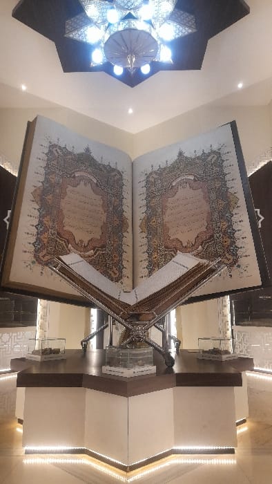 An expansive, open replica of the Quran is centrally displayed on a multi-faceted platform within an exhibition hall located in Glantengan, Indonesia. The colossal book, positioned on a polished metallic stand, features two visible pages with intricate, multi-colored Islamic calligraphic and geometric borders surrounding Arabic script. The platform, constructed with a dark wooden top and a light-colored base, is illuminated by white LED strip lighting along its lower edge. Two smaller glass display cases containing indiscernible artifacts are situated on the platform. The ceiling, incorporating a dark star-shaped recess, features a large, ornate chandelier with multiple bright lights and additional recessed spotlights. The surrounding environment consists of light-colored walls, some with subtle geometric patterns. No individuals are present within the frame.