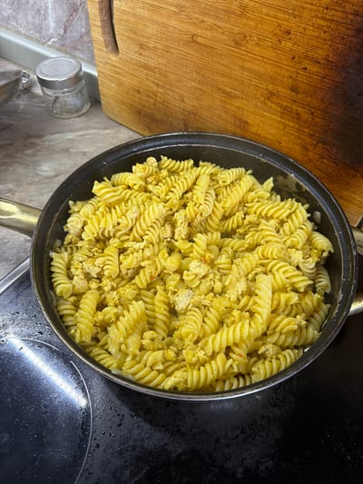 This overhead shot captures a portion of fusilli pasta mixed with what appears to be crumbled scrambled eggs in a black frying pan with a gold handle. The pasta is a bright yellow, indicating it may be cooked with turmeric or saffron. The scrambled eggs are also yellow and speckled with what could be herbs or spices. The pan sits on a black stovetop, which has visible water droplets on its surface. To the left of the pan is a wooden cutting board with a textured surface, suggesting it is well-used. The cutting board has a rounded edge and a visible handle or groove. Below the cutting board and to the left of the pan, a small glass jar with a silver lid is visible, sitting on a marbled countertop. The overall scene suggests a domestic kitchen setting, likely in the morning or during meal preparation. No people are visible in the image. The lighting appears to be artificial, common in indoor environments. There is no visible text.