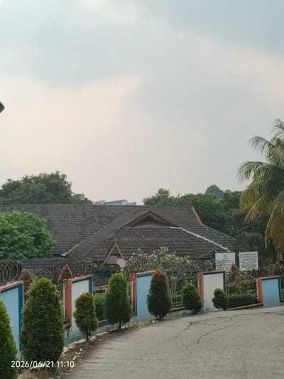 The image shows a residential building with a dark tiled roof, surrounded by lush green trees and a tall palm tree on the right. A blue and orange fence runs along the property line, with several neatly trimmed conical evergreen trees planted in front of it. There are also some thorny wires on top of the fence. In the background, a modern building with a blue facade is visible. The sky is overcast. A sign on the fence reads "RAUDHATUL ATHIFAL" and "TERAKREDITASI A". The date stamp at the bottom left corner of the image indicates "2026/06/21 11:10". The scene is a residential area in Bojong Gede, Indonesia.