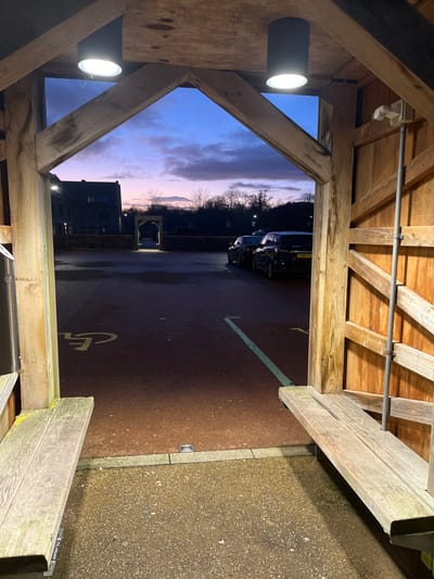 The image depicts an outdoor scene at twilight, viewed from within a weathered wooden-framed structure in Newton St. Loe, United Kingdom. The foreground is defined by robust timber posts and beams forming an A-frame aperture. Two black cylindrical spotlights, actively illuminated, are mounted on the overhead beams. Integrated into the structure on both the left and right are weathered wooden benches. A light gray aggregate surface constitutes the ground directly beneath the viewpoint.
Beyond the wooden structure, a dark reddish-brown tarmac parking area is visible. A yellow disabled parking symbol is painted on the asphalt to the left. On the right, a faded green line marks a boundary. Two dark-colored vehicles are parked on the right side of the tarmac; the closer vehicle is a dark SUV with license plate 'FY66 VTF'. In the midground, a low stone wall or hedge delineates the parking area, with a stone archway visible directly ahead. A multi-story building with dark exterior walls is present on the far left, situated behind a stone wall. The distant horizon is characterized by bare trees and dark vegetation silhouetted against a twilight sky, which displays a gradient from deep blue overhead to shades of pink and purple near the horizon, with scattered clouds. No individuals are present.