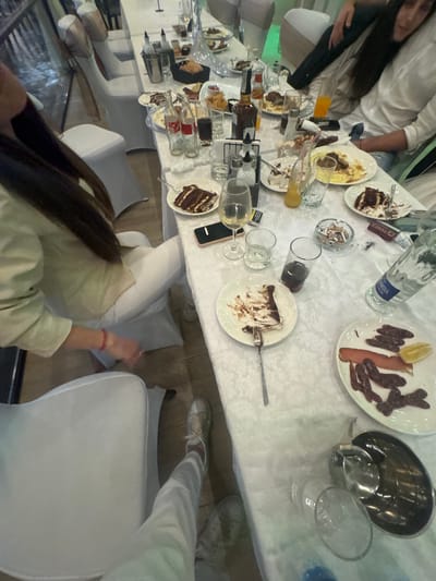 The scene depicts a table covered with a white tablecloth, likely in a restaurant or event space in Kovachite, Bulgaria. Several people are seated around the table, though only their arms and parts of their bodies are visible. There is an assortment of food and drinks on the table, including plates with partially eaten cakes, sliced sausages, a bottle of water labeled "SANDANSKI", glasses, and silverware. Some glasses appear empty, while others contain liquids. The table shows signs of a meal in progress. There are also mobile phones, possibly in use. White chairs surround the table. The image captures a candid moment during what seems to be a social gathering.