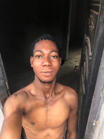 A young adult male, appearing to be of Black ethnicity, is depicted from the mid-torso upward, centered in a doorway within the city of Oraukwu, Nigeria. He has dark, short, textured hair styled with small braids or twists, a light beard on his chin, and full lips. His expression is a slight smile, and he gazes directly at the camera. He is shirtless, displaying his bare chest and shoulders, and wears a gold-colored chain necklace with a gold-colored cross pendant. The background consists of a dimly lit interior space, likely a room or hallway, with a dark, textured door frame or wall visible immediately to the viewer's right. Further back, another dark doorway or opening leads to a deeper, unlit area. The lighting suggests an interior setting with ambient light originating from the direction of the camera.