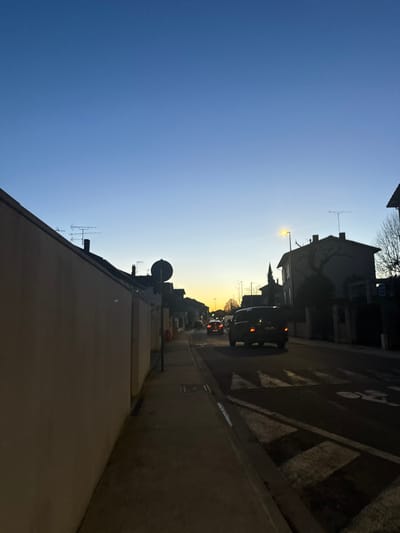 This image depicts a residential street in Albi, France, at dusk. The sky transitions from a deep blue overhead to a lighter blue and then to an orange-yellow glow on the western horizon, indicating sunset. Artificial lighting is active.
In the foreground on the left, a light-colored, smooth wall, likely stucco or concrete, runs diagonally from the lower left towards the midground. The lower portion of this wall is in shadow. Adjacent to the wall is a concrete sidewalk, extending into the midground.
The street, positioned to the right of the sidewalk, features white painted road markings, including a speed bump or traffic calming device with alternating dark and light triangular patterns, and a partial white bicycle symbol.
Several vehicles are present on the asphalt road, all facing away from the viewer, moving towards the receding sunset. The closest vehicle is a dark-colored minivan or SUV, its red taillights illuminated. Behind it, at least two additional vehicles with illuminated red taillights are visible further down the street.
Along the street, houses and residential buildings are silhouetted against the twilight sky. On the left side of the street beyond the wall, rooflines with television antennas and chimneys are discernible. A dark, circular sign on a pole stands on the sidewalk. On the right side of the street, a multi-story building with a distinctive roof and a bare-branched tree in front of it is visible. A streetlamp with a yellow light source is illuminated, casting a glow onto the road and adjacent structures. The overall scene suggests a quiet, suburban environment as evening approaches.