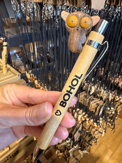 An adult's light-skinned hand holds a light-tan bamboo pen in the foreground. The pen features a silver-colored metal clip and tip, and is inscribed with a black heart symbol followed by the word "BOHOL" in black capital letters. In the midground, directly behind the pen, hangs a wooden charm shaped like a Tarsier head, distinguished by large yellow-orange eyes. The background consists of numerous black-corded necklaces or keychains with small charms, suspended from a dark display rack. A second, partially visible bamboo pen, also marked "BOHOL," rests on a display stand to the left. The scene depicts a souvenir shop in Panglao, Philippines.