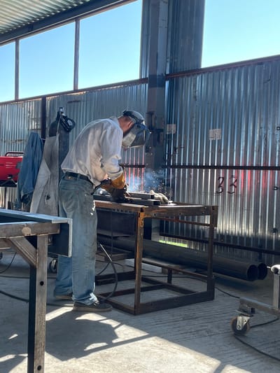 A person is welding metal in an industrial workshop. The individual is wearing a white long-sleeved shirt, blue jeans, welding gloves, and a welding helmet. Sparks are visible from the welding process. In the background, there are metal walls, pipes, and a welding machine. The floor is concrete, and sunlight is streaming in from a large window. The scene appears to be taking place in the city of Sokury, Russia.