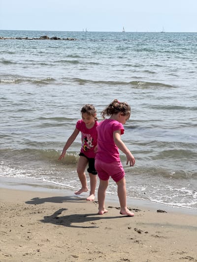 Two girls are playing on a sandy beach next to the sea. The girl on top is wearing a pink outfit, and the one underneath is wearing a dark shirt with a Minnie Mouse design and dark shorts. They are standing on the edge where the sea meets the sand, their feet in the water. The water is slightly choppy, and the sky is light. The location is Nettuno, Italy.