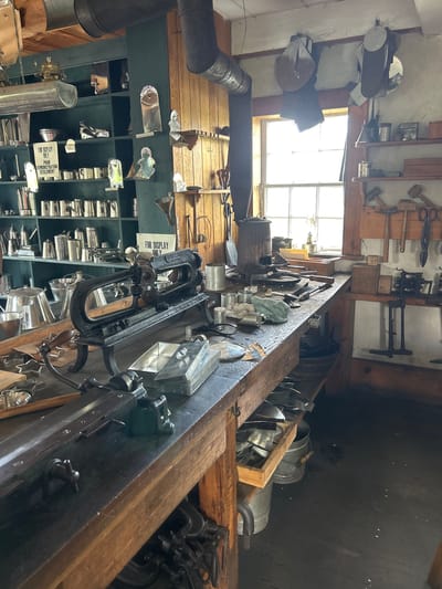 This is a photograph taken inside what appears to be a workshop or historical display, possibly a tin shop, given the prevalence of metal items. The scene is filled with various metal objects, including buckets, cones, tins, and what look like kitchenware or industrial tools. The items are displayed on shelves and on a long wooden workbench that runs through the center of the image.

The workshop setting is evident from the tools hanging on the wall, such as hammers, scissors, and other metalworking implements. A large window with multiple panes allows natural light to illuminate the space, suggesting it is daytime. The overall impression is one of a historical or artisanal setting, possibly a museum or a preserved workshop.

There are no people visible in the photograph. The lighting suggests it is daytime, and the interior setting implies no specific weather conditions are relevant. Location cues are minimal, but the presence of tinware and traditional tools might suggest a historical craft.

One visible sign on a shelf reads "FOR DISPLAY ONLY POUR DEMONSTRATION SEULEMENT," which translates to "For Display Only Pour Demonstration Only." This further supports the idea of a museum or educational display. The image is oriented sideways, with the workbench running horizontally across the frame and the shelves vertically. The focus is on the objects and the workshop environment, creating a sense of a bygone era of craftsmanship.