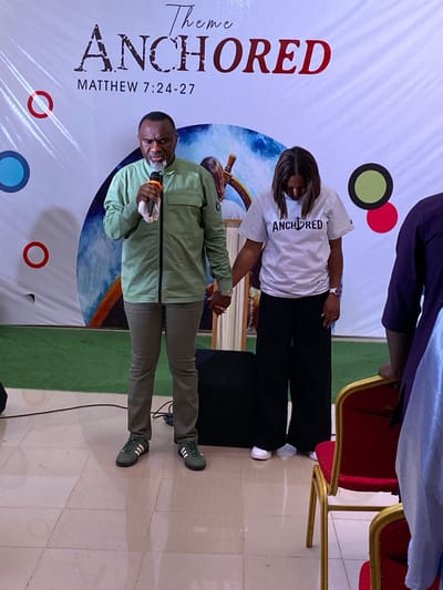 A sideways shot taken indoors shows a man and a woman on a stage standing in front of a banner. The banner is white with a graphic of a ship's wheel. The words "Theme ANCHORED MATTHEW 7:24-27" appear vertically on the left side of the banner. The man wears a light green shirt, pants, and sneakers. He holds a microphone to his mouth with his right hand. His left hand is extended, and he appears to be holding the woman's right hand. She is wearing a white t-shirt, black pants, and white shoes. The word "ANCHORED" is printed across the front of her t-shirt. A black speaker sits behind her. There are two red and gold chairs to the right of the woman. A section of a dark purple robe can be seen above the woman's head. A green area can be seen on the right side of the shot. The location is a stage in the city of Miango, Nigeria.