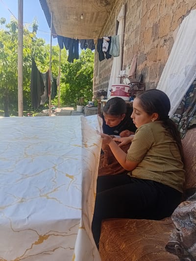 Two young girls are seated at a table covered with a white tablecloth patterned with gold marbling. The girl on the right, wearing a green short-sleeved shirt and black leggings, is leaning forward, her left hand resting on the tablecloth. The girl on the left, wearing a black t-shirt, is also looking at the tablecloth. The background features a rough stone wall, laundry hanging on a clothesline, lush green trees, and a clear blue sky. The scene is set outdoors, possibly in a rural area, consistent with the village of Karbi, Armenia, known for its traditional architecture and surrounding natural landscapes.