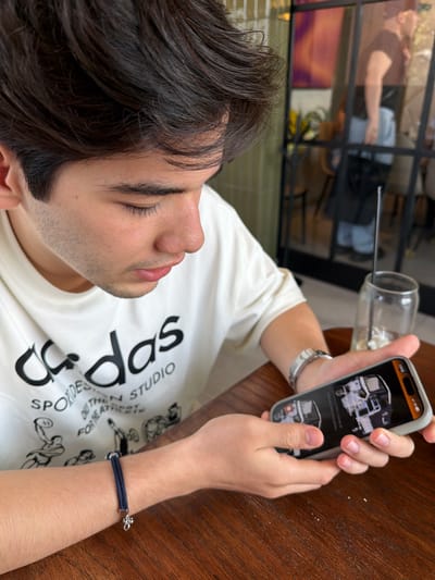 A young male, with dark hair and light facial stubble, is depicted indoors at a wooden table in Veracruz City, Mexico. He wears an off-white t-shirt with a black "Adidas SPORTDESIGN STUDIO" logo and graphics. On his right wrist is a silver-toned watch, and on his left, a dark blue braided bracelet with a silver-toned charm. The male holds a light-cased smartphone horizontally with both hands, viewing content on its screen. The screen displays what appears to be a black-and-white image of a vehicle's interior or a technical schematic. To the right, on the table, a clear glass jar containing a dark straw is present. The background features a large, dark-framed glass partition, which reflects the partial image of an individual, likely male, wearing a dark shirt and light pants, holding a dark bag while standing. The overall environment suggests a casual commercial establishment such as a cafe or restaurant.