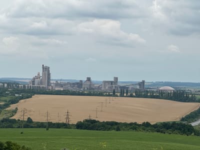 A wide-angle, elevated shot captures a rural industrial landscape under a cloudy sky. In the background, a large cement plant complex dominates the horizon, characterized by tall concrete silos, conveyor belts, and various industrial structures. To the right of the plant, a large, domed building is visible. In the mid-ground, a vast, dry, golden-brown field stretches across the frame, marked by tire tracks from agricultural machinery and traversed by a line of electrical transmission towers. A band of dark green trees separates the dry field from a lower foreground of vibrant green fields. The scene is devoid of any human presence. The overall impression is one of a working industrial area integrated into a predominantly agricultural environment.