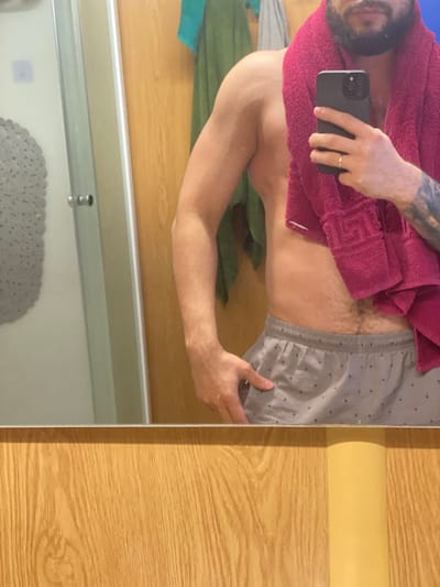 In a bathroom, a person is taking a mirror selfie. They are wearing grey patterned shorts and a pink towel is draped over their shoulder and chest. The person is holding a black smartphone in their right hand, with a gold ring on their left ring finger. The background shows a wooden door, a towel hanging on a hook, and a shower curtain. The location is an unknown city in Bangladesh.
