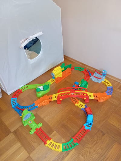 A child's colorful plastic train track set is laid out on a light brown parquet floor. The track is arranged in a winding path, incorporating various colored pieces including red, yellow, blue, and green. The set features several accessories such as bridges, a station, and a ramp. A white play tent, partially obscuring the background, is positioned to the left of the train set. The tent has a circular opening with a checkered cloth draped over it. Inside the tent, pillows in shades of blue and green are visible, along with a beige stuffed animal. The lighting suggests an indoor environment, possibly a child's room or play area. The scene does not provide any specific information about the city of Baden, Austria.