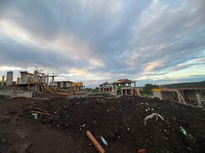 The image depicts a construction site in Cherni Vrah, Bulgaria. Several unfinished concrete structures, likely houses, stand on uneven ground. The foreground is dominated by piles of dark soil littered with construction debris, plastic bottles, and other trash. Wooden planks and formwork are scattered around the site. The sky is overcast with a mix of grey and white clouds, and there is a hint of light on the horizon. No people are visible in the image.