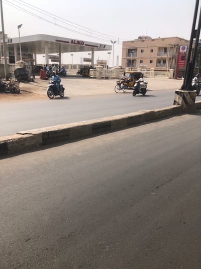The scene depicts a daytime street view in Gwammaja, Nigeria. The foreground consists of an asphalt roadway bordered by a low concrete curb, painted with alternating black and light-colored stripes, separating it from the street level further back.

In the mid-ground, multiple individuals on various vehicles are in motion. From left to right: a person in a light blue garment operates a dark motorcycle, moving rightward. Adjacent to this, a light blue bicycle is ridden rightward by an individual wearing a light-colored top. Immediately to its right, a person in a light-colored top drives a light blue scooter or moped, also traveling rightward. A fourth motorcycle, partially obscured by a dark, striped concrete pillar on the far right, is also moving rightward.

The background features an "ALIKO" brand gas station on the left, characterized by a prominent white canopy with red lettering and visible fuel pumps. Several individuals are present within the gas station compound, interacting near parked motorcycles and yellow auto-rickshaws. A low, decorative white fence separates the gas station from the street. To the right of the gas station stands a multi-story, light-colored building with multiple windows and balconies, displaying a red and white sign on its right facade. Overhead utility lines traverse the clear, pale sky. A pile of debris and a dark utility cart are visible near the left entrance of the gas station.