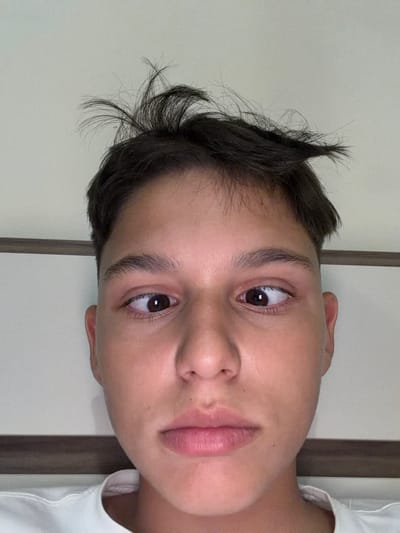 A young individual, appearing to be male, is depicted in a close-up, head-and-shoulders portrait, likely taken indoors in Yoakim Gruevo, Bulgaria. The subject has short, dark brown hair that is noticeably dishevelled and spiked upwards on top. Their skin tone is light, and they possess dark brown eyes that are purposefully crossed, creating an esotropia effect. The nose is prominent, and the lips are full with a neutral, slightly downward turn. The subject is wearing a white crew-neck t-shirt, a portion of which is visible around the neck and shoulders. The background consists of a plain, light-colored wall with a horizontal dark brown wooden trim or panel running across the mid-section behind the subject's head and shoulders. Lighting appears bright and even, illuminating the subject from the front.