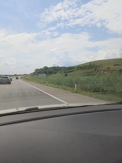 The image shows a view from inside a car, looking out at a highway on a cloudy day. The road curves to the left, with several cars visible in the distance. To the right of the road, there is a grassy hill covered in trees and vegetation. The sky is filled with white, fluffy clouds against a light blue background. The foreground of the image shows the dashboard of the car. The scene is a typical highway landscape, likely in or around Mineralnye Vody, Russia, given the vegetation and road infrastructure.