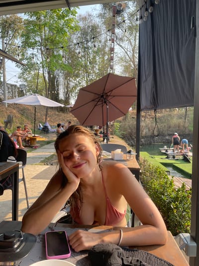 A young woman is reclining at a table in an outdoor setting at a location in Tambon Nam Phrae, Thailand. She is wearing a swimsuit and resting her head on her hand. A mobile phone is lying next to her on the table. An outdoor seating area with umbrellas and other patrons can be seen in the background. A body of water with people participating in water activities is also visible in the distance.