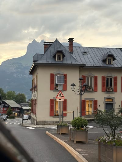 The image depicts a street scene in Saint-Gervais-les-Bains, France, with a multi-story building featuring red shutters and a gray metal roof in the foreground. A prominent mountain looms in the background under a cloudy sky. The building has several windows, some adorned with flower boxes. A street lamp stands near the building, and a pedestrian crossing sign is visible. The street itself is wet, suggesting recent rain, and there are planters with vegetation along the sidewalk. Other buildings and parked vehicles are visible further down the street.