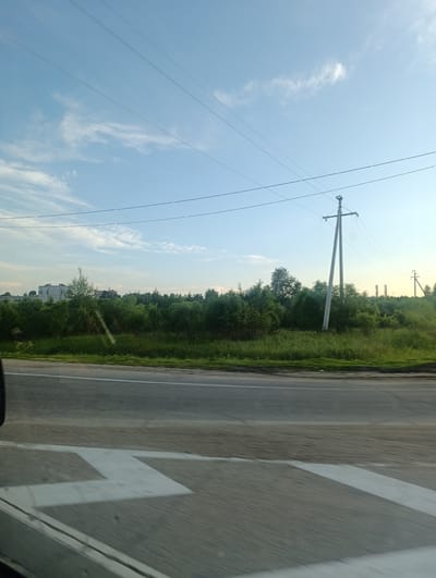 The image depicts a roadside scene with a clear blue sky dotted with wispy clouds. Power lines stretch across the sky, supported by a tall utility pole in the mid-ground. To the left, a low-rise building or complex is visible behind a dense line of green trees and bushes. The vegetation appears lush, suggesting a temperate climate. In the foreground, a paved road is visible with white lane markings, including a chevron pattern indicating a lane split or merge. The perspective suggests the photo was taken from a moving vehicle. The overall impression is one of a rural or semi-urban landscape on a bright day. There is no visible text in the image.