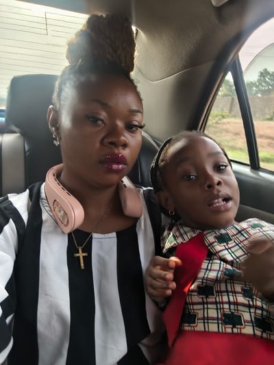 A medium shot captures a woman and child in a car, likely a sedan, based on the interior. The woman is positioned on the left, wearing a black and white striped shirt, a gold cross necklace, and a light peach-colored portable neck fan. Her hair is styled in a high bun. The child is seated next to her on the right. The child is wearing a red and white patterned shirt. The child has small gold hoops in their ears. The background reveals the car's interior, with glimpses of trees and greenery visible through the window, indicating they are likely traveling through a suburban or rural area. The location is stated as Nabatum, Nigeria. The interaction between the subjects is apparent in their proximity, though their specific actions or conversation are not discernible from the image.