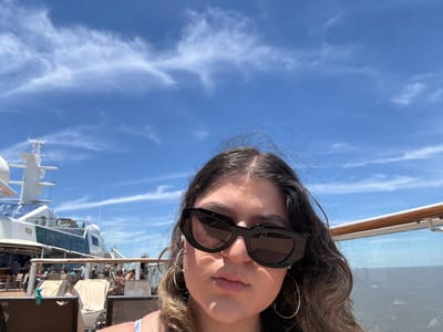A person with long brown hair and wearing black sunglasses and large hoop earrings poses against a bright blue sky dotted with wispy white clouds. Part of a cruise ship is visible in the background, including the railing, lounge seating, and the ship's communication tower and equipment. The image appears to have been taken outdoors on a sunny day.